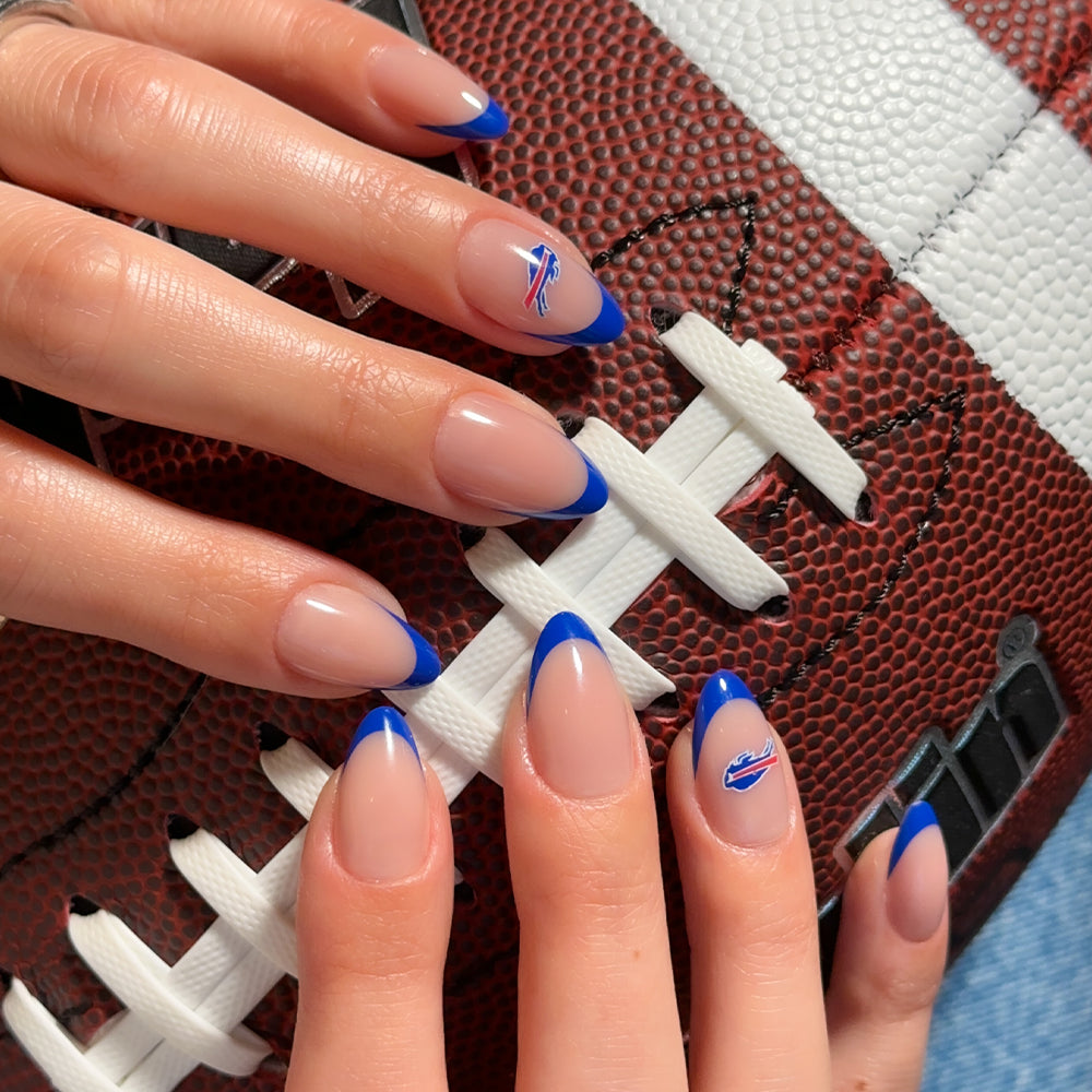 Hands with almond-shaped nails wearing glamnetic NFL Press-On Nails, featuring blue French tips and a small football team logo on two nails, holding a brown leather football with white laces. - Product image