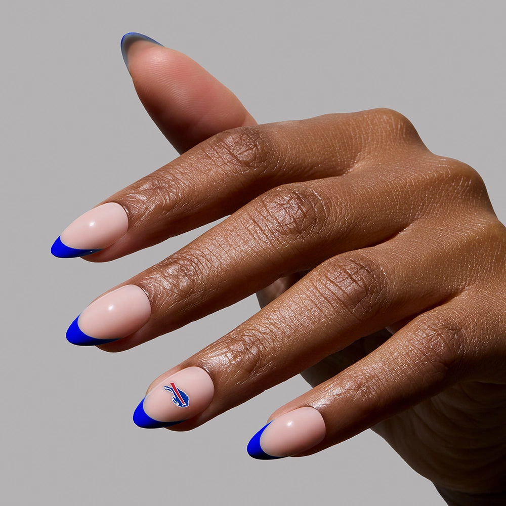 A hand with almond-shaped nails painted nude with blue French tips wears the glamnetic Buffalo Bills Press-On Nail Set, featuring a small red, white, and blue team logo accent. The background is plain gray. - Product image