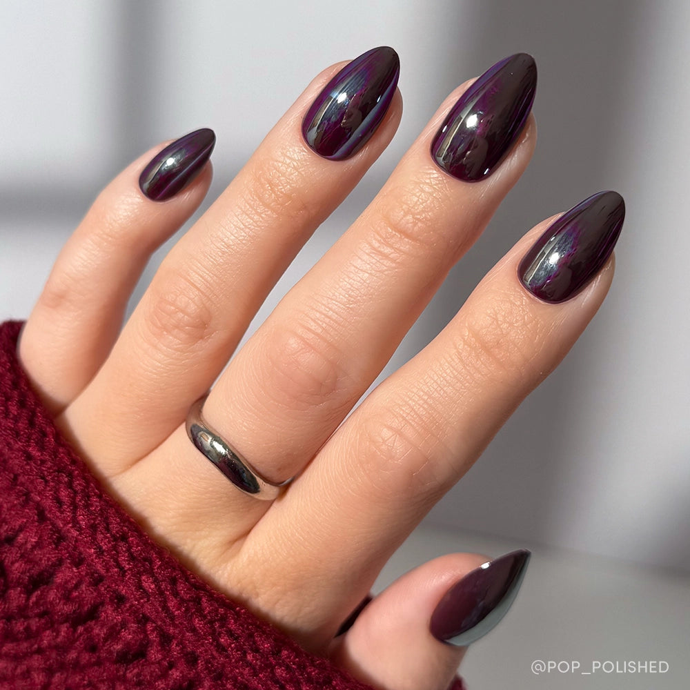 A hand with short almond-shaped nails painted in shiny, dark purple Burgundy Chrome polish by glamnetic. A silver ring and a textured dark red sweater sleeve complete the chic look of this burgundy chrome nail set. - Product image