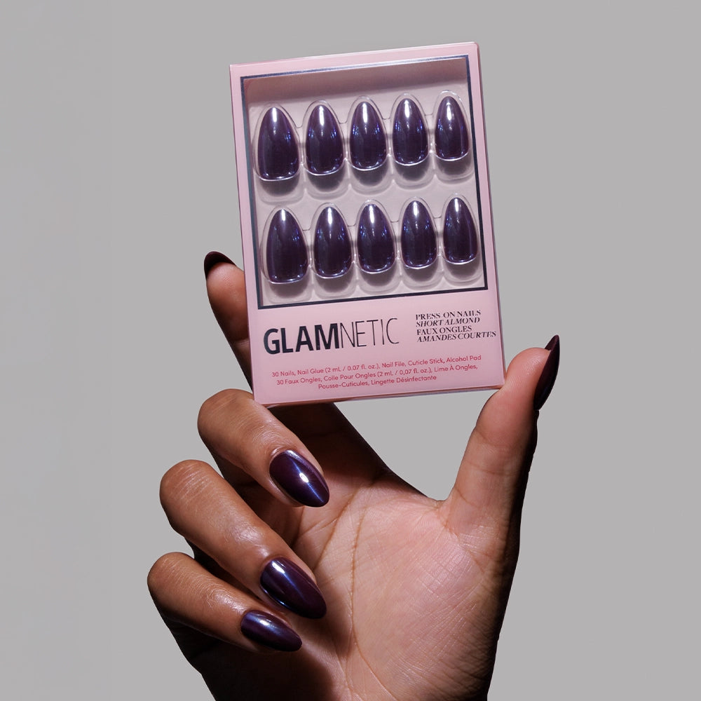 A hand with dark purple, glossy nails holds a glamnetic Burgundy Chrome press-on nail box, featuring ten reusable nails in two rows, set against a plain gray background.