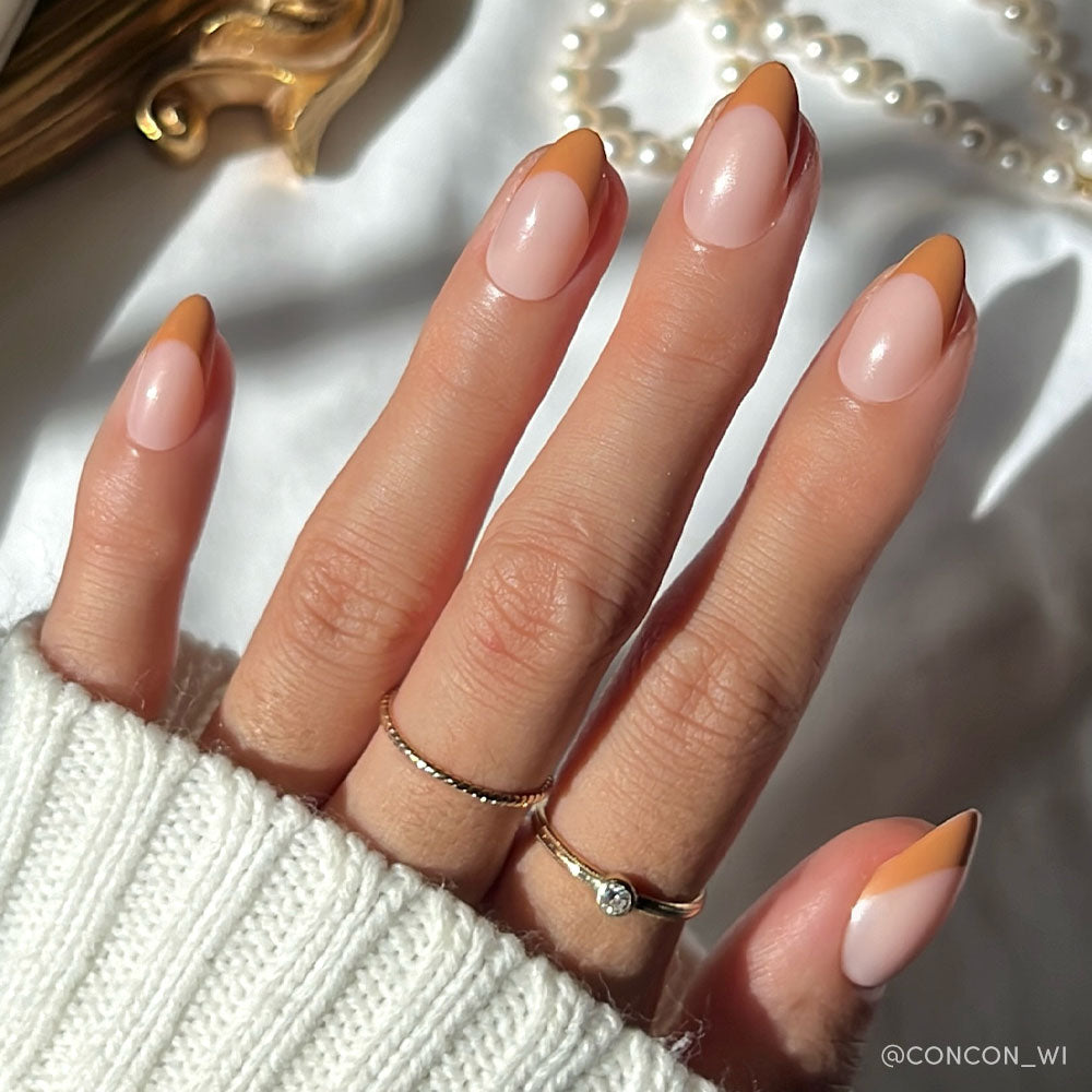 A hand with almond-shaped nails in glamnetic’s Café Crème, showcasing reusable peachy-orange French tips. Two gold rings and a white knit sweater complete the cozy look, with pearls softly blurred in the sunlit background. - Product image