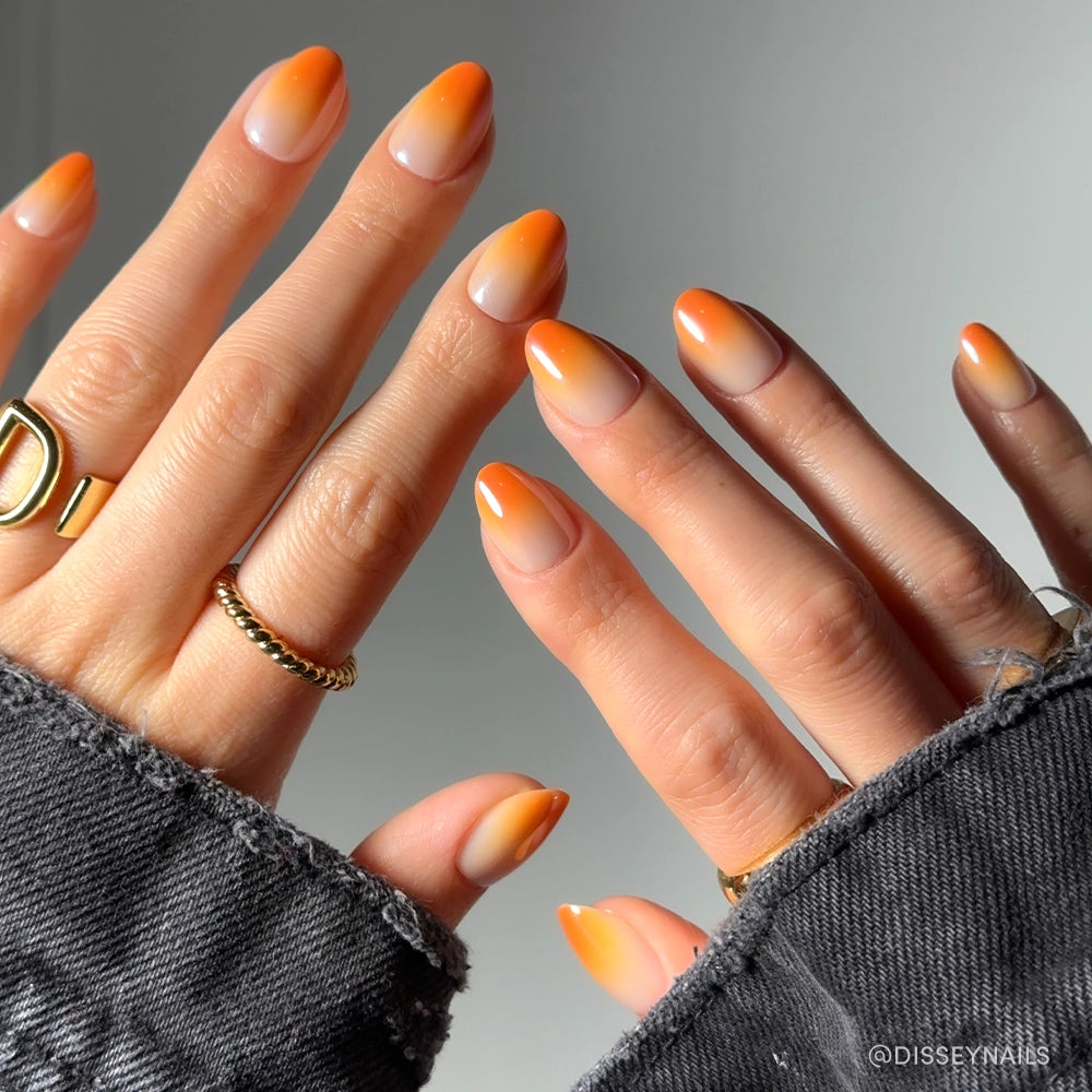 Close-up of hands with almond-shaped glamnetic Candy Corn nails, showcasing bright orange French tips and a subtle ombré effect. Gold rings and a frayed dark denim jacket complete the festive Halloween look. - Product image