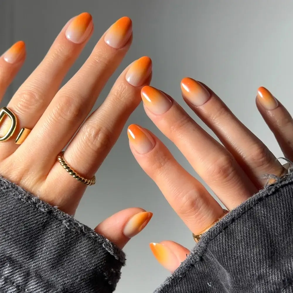 Two hands with short oval nails display the Candy Corn nail set by glamnetic, featuring an orange gradient design inspired by classic candy corn. The hands are adorned with gold rings, and the person is wearing a black denim jacket against a softly blurred neutral background. - Product image