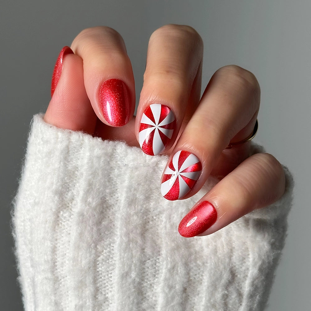 A hand adorned with glamneticās Candy Crush press-on nailsāfeaturing red glittery nails and two accent nails with red and white pinwheel designsāpeeks from beneath a white knit sleeve, displaying the festive short oval shape thatās perfect for holiday-ready manicures. - Product image
