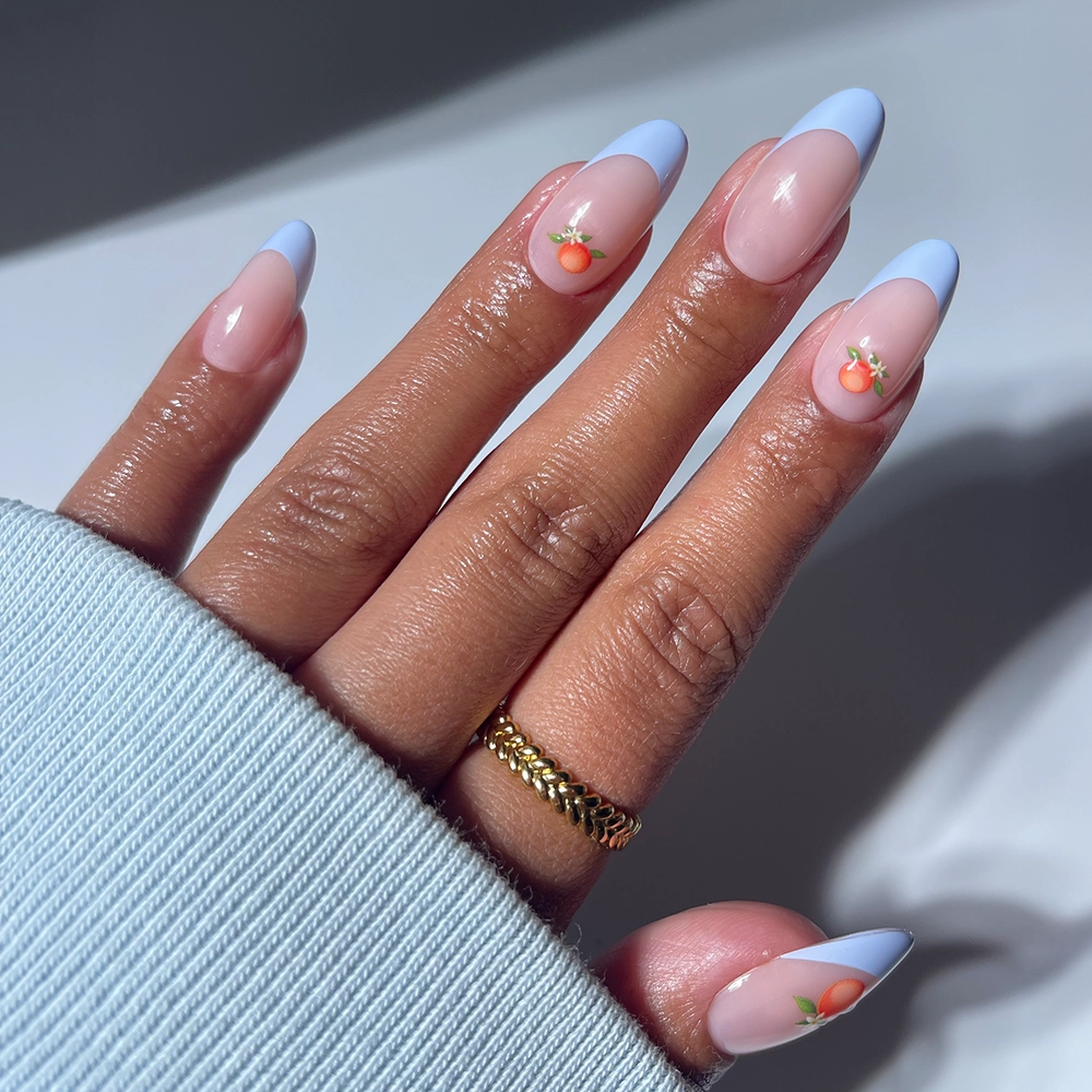 A hand with medium oval Capri Coast nails by glamnetic, featuring pink and blue French tips with peach designs, is shown. The person wears a gold ring and a light blue sweater, embodying the playful vibe of Capri Coast.