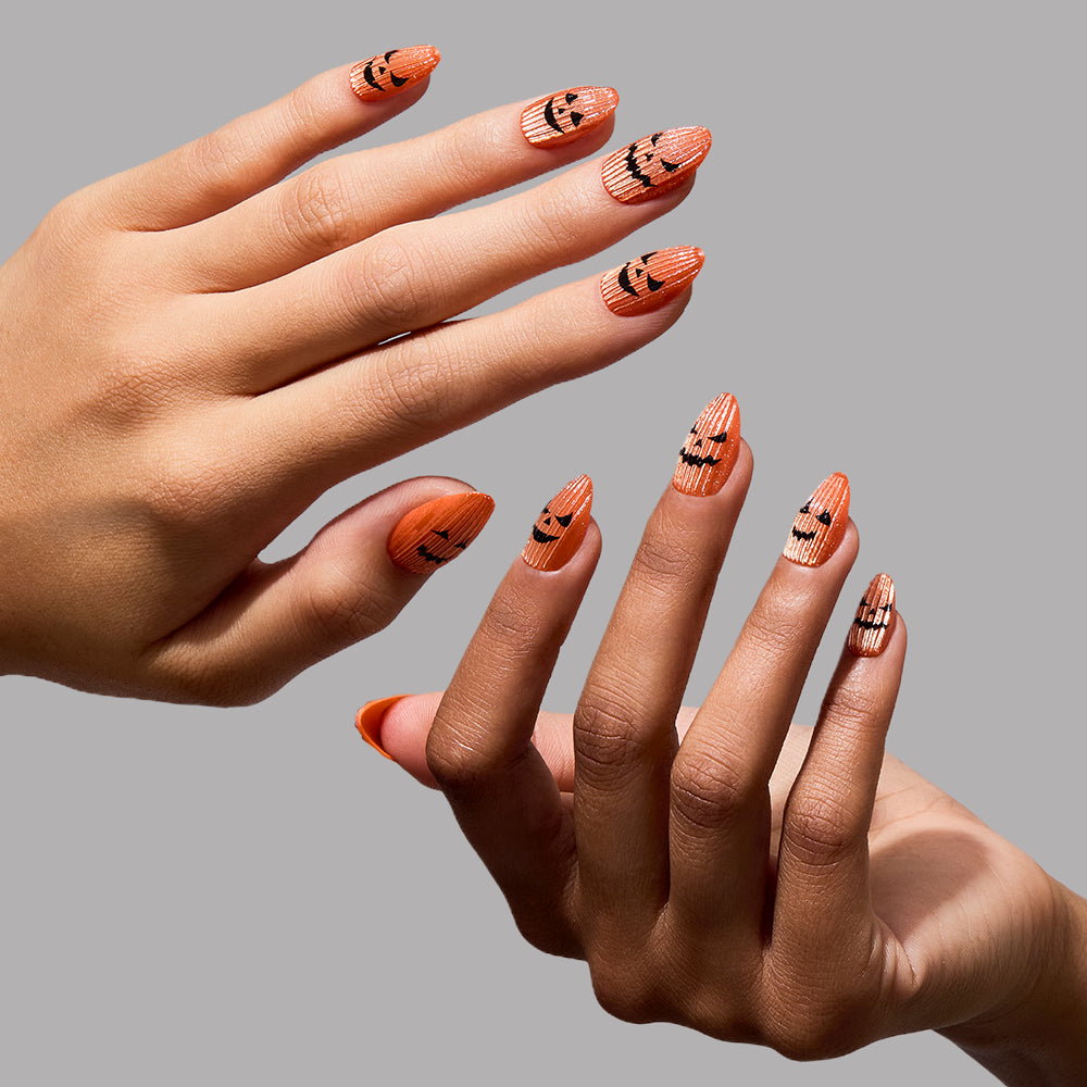 Two hands display glamnetic’s Carving Contest press-on nails—long, ribbed orange nails each featuring black Jack-o-lantern faces in a playful pumpkin nail art style—against a plain gray background. - Product image