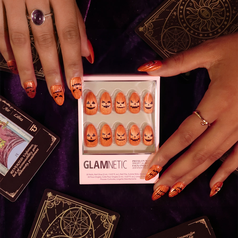 Hands adorned with Carving Contest metallic orange jack-o-lantern press-on nails from glamnetic hold a branded box featuring the same pumpkin nail art. The scene is set against a backdrop of rich purple velvet scattered with illustrated tarot cards, perfectly accentuating the playful velvet finish of these festive nails for an enchanting, bewitching vibe. - Product image