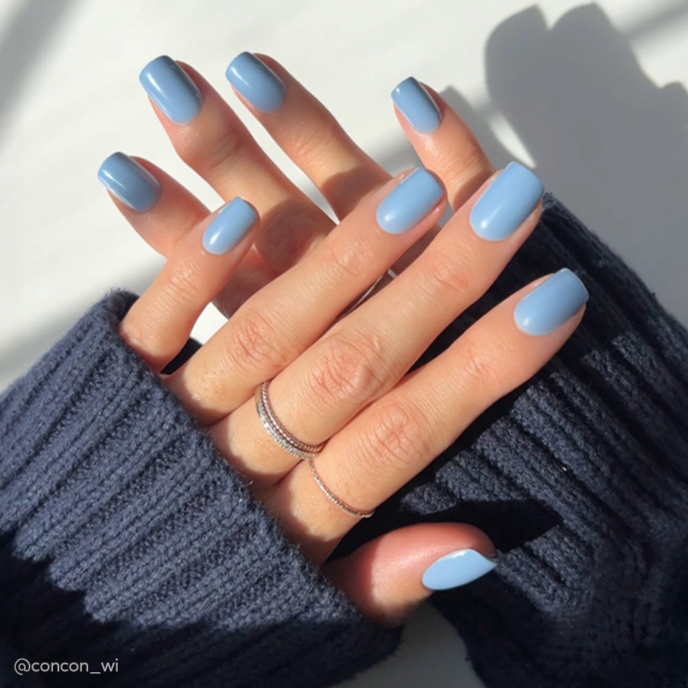 Two hands with neatly manicured short squoval nails painted in glamnetic’s Cashmere Blue rest on each other. The person wears a navy blue sweater and thin silver rings, while sunlight casts soft shadows on a light background. - Product image