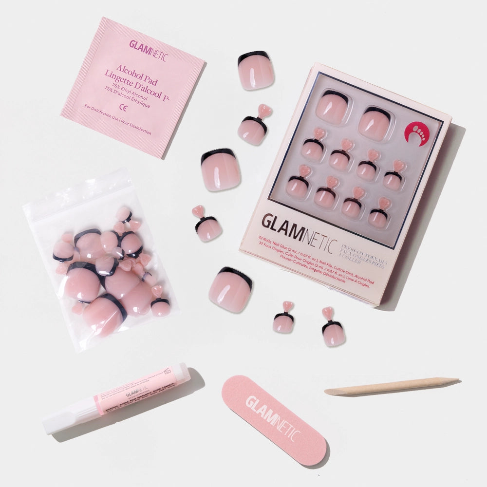 A flat-lay of the Glamnetic nude pink press-on toenail kit with bold black French tips, including glue, file, stick, and alcohol pad. - Product image