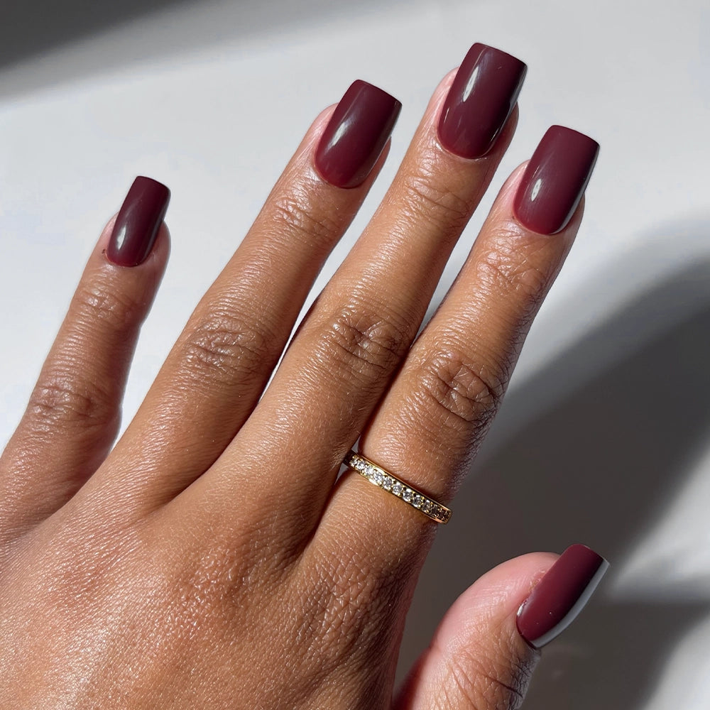 A hand with medium square Cherry Mocha nails by glamnetic in a cherry brown gloss is shown with a gold chain ring on the middle finger, set against a softly blurred background. Ideal for fans of vegan, cruelty-free style. - Product image