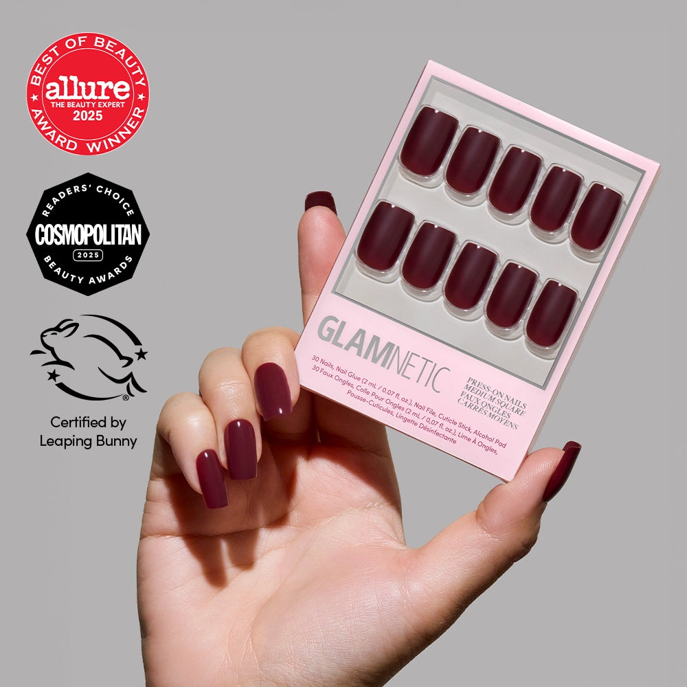 A hand with vegan, cruelty-free maroon Cherry Mocha press-on nails holds a pink glamnetic box containing matching medium square nails, all set against a plain light grey background. - Product image