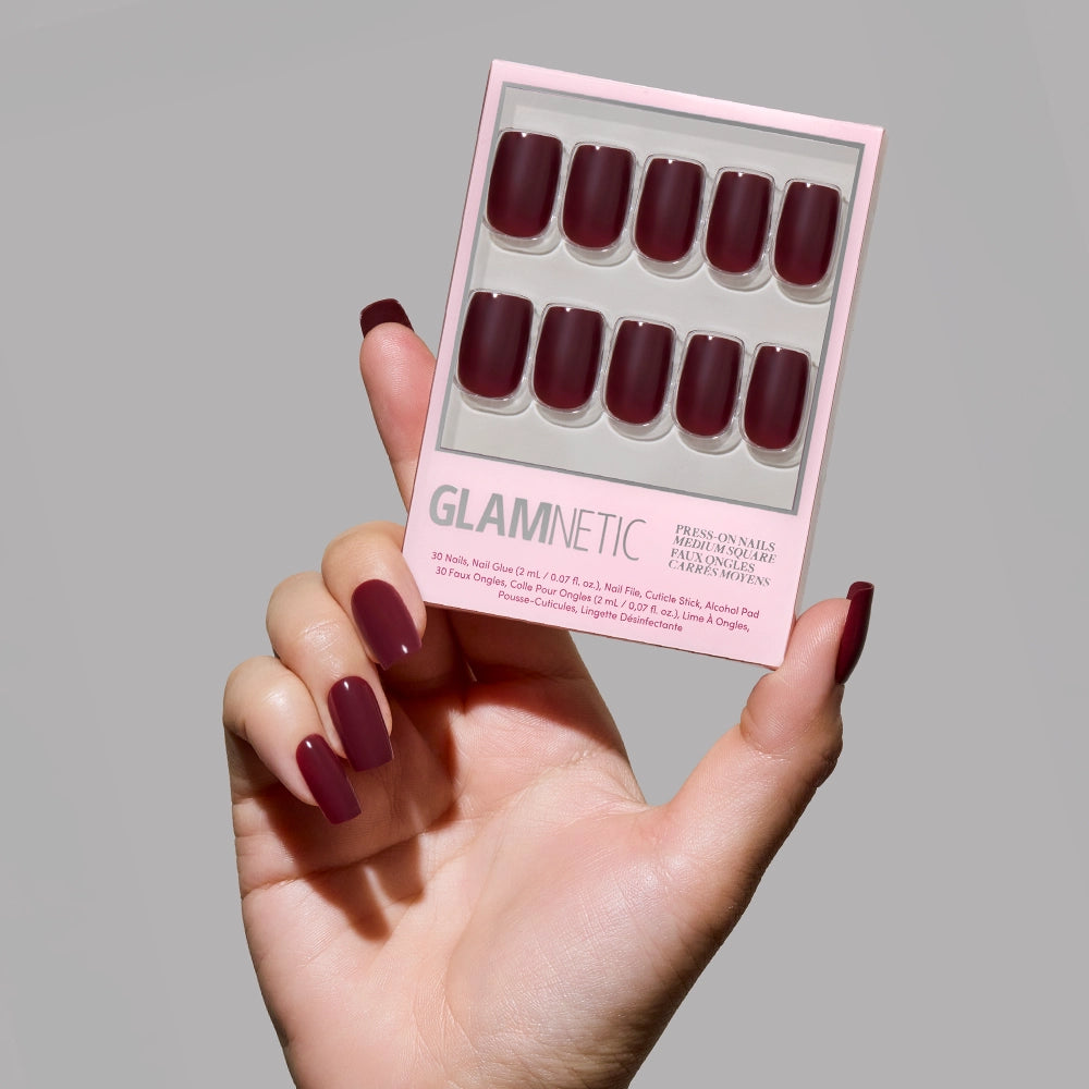 A hand with vegan, cruelty-free maroon Cherry Mocha press-on nails holds a pink glamnetic box containing matching medium square nails, all set against a plain light grey background.
