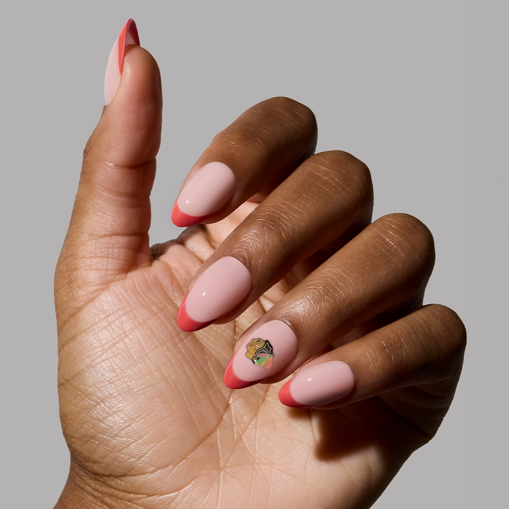 A hand wears the glamnetic Chicago Blackhawks Press-On Nail Set, featuring almond-shaped nails with a pink base, bright pink fandom french tips, and a butterfly accent on the ring finger, displayed against a plain gray background. - Product image