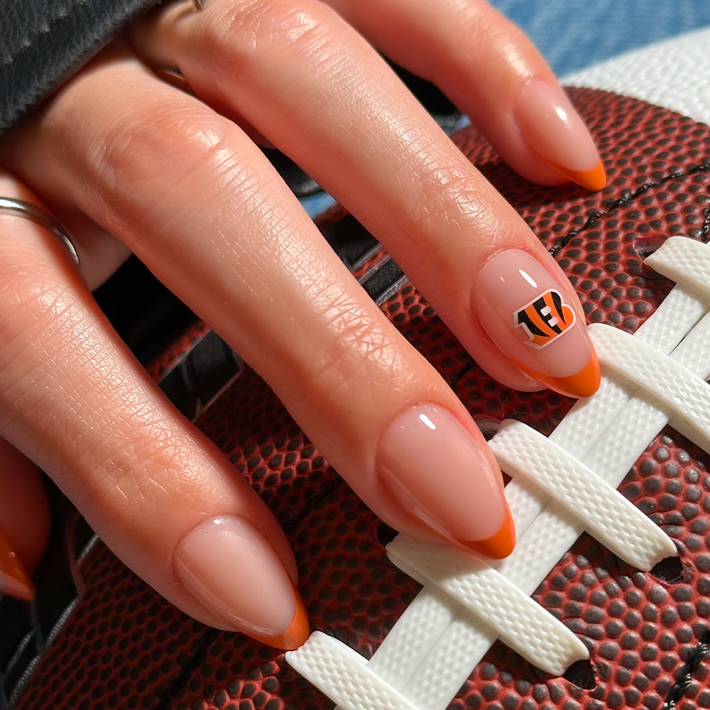 A hand wearing glamnetic NFL Press-On Nails, featuring orange French tips and a Cincinnati Bengals logo accent nail, rests on a textured football with white laces. - Product image