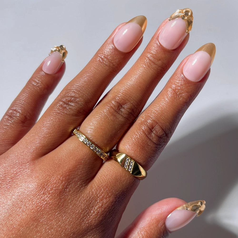 A hand with almond-shaped nails, showcasing glamnetic's "Citrine" design with gold foil French tips and decorative accents, wears two gold rings with embedded stones against a neutral background.