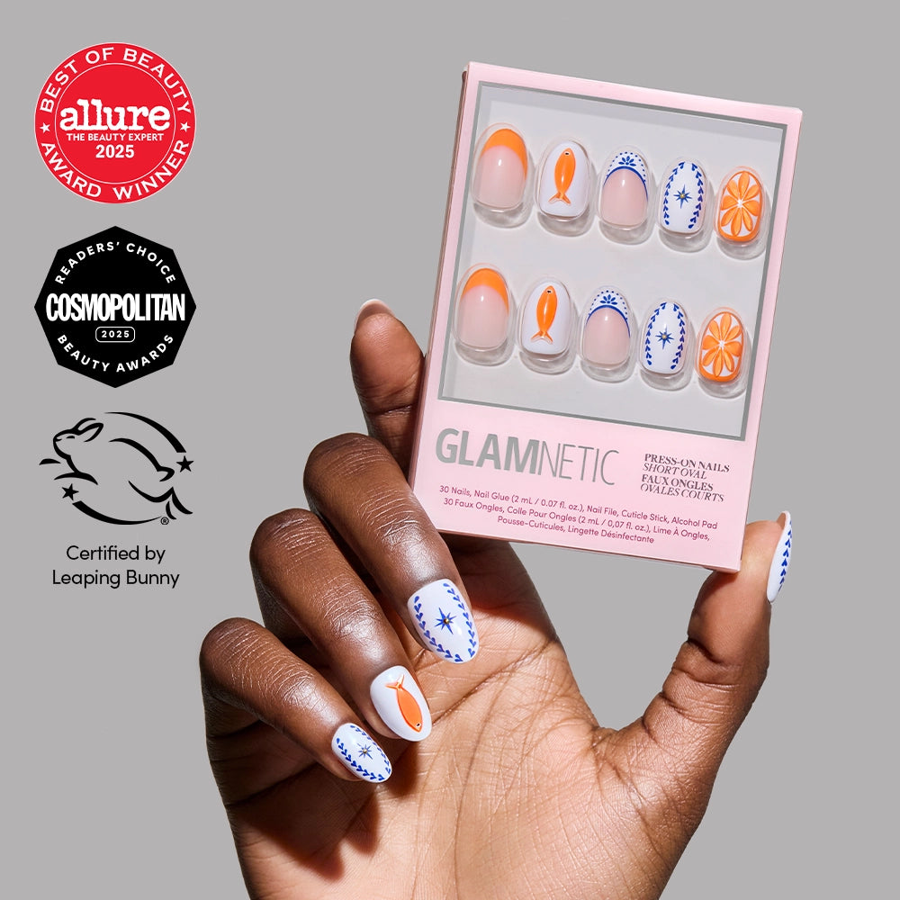 A hand with decorated short oval nails holds a box of glamnetic Citrus Coast Press-On Nails with carrot and blue floral designs. Allure, Cosmopolitan, and Leaping Bunny award logos are displayed on a gray background.