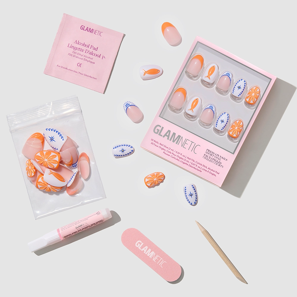 The glamnetic Citrus Coast set includes short oval press-on nails with orange slice and blue leaf designs, extra nails, a pink nail file, wooden cuticle stick, glue tube, and alcohol pad—all arranged on a light background. - Product image