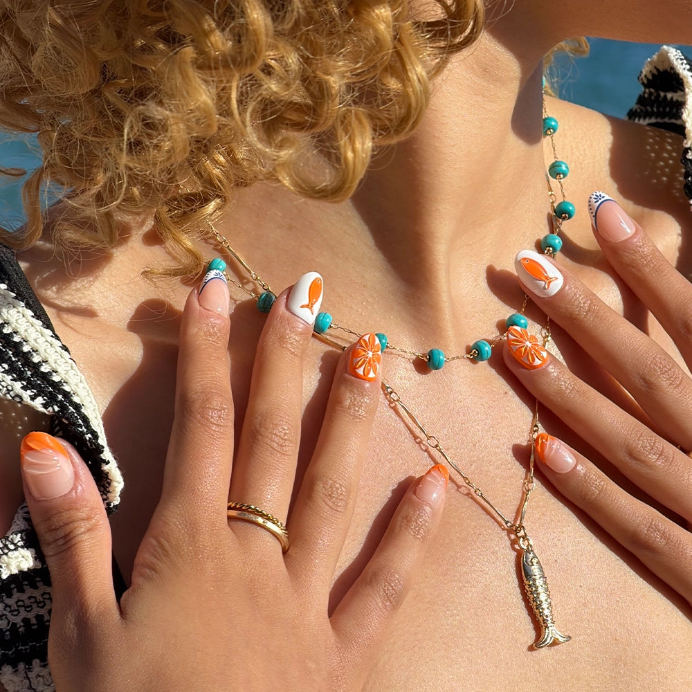 A woman with curly blonde hair shows off her glamnetic Citrus Coast press-on nails, plus a turquoise beaded necklace and gold fish pendant, holding her hands near her chest in bright sunlight for a salon-quality manicure. - Product image