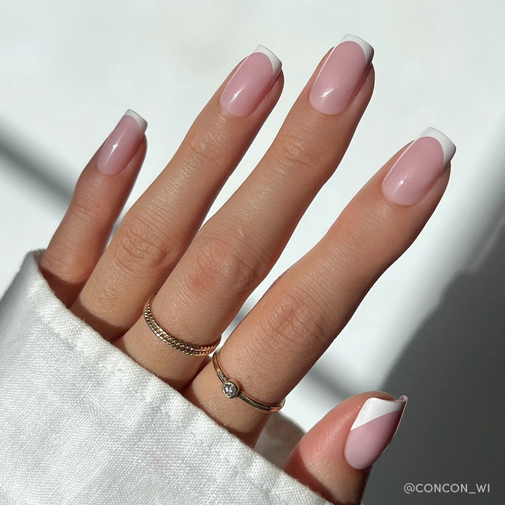 A hand with long, almond-shaped nails flaunts glamnetics Classy French mani: a modern twist on French tips featuring a light pink base and white geometric accents. Gold rings and a white cuffed sleeve complete the look. - Product image