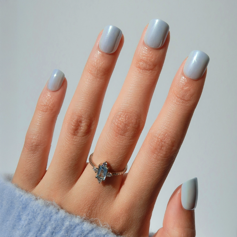 A hand with short nails painted in glamnetic's Clear Skies, a light blue glossy polish, wears a silver ring with an oval gemstone. The wrist is partially covered by a fuzzy light blue sleeve, shown against a plain background.