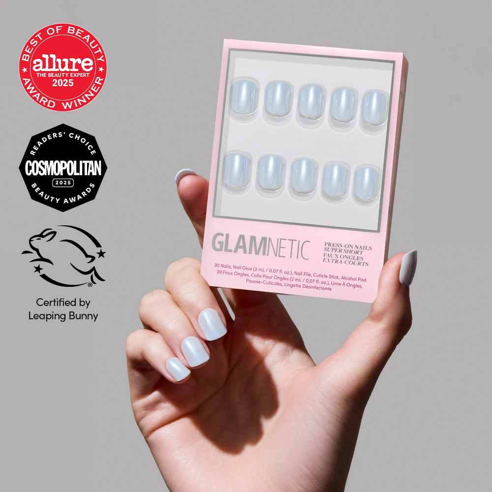 A hand with iridescent blue salon-quality Clear Skies press-on nails holds a glamnetic nail kit. The kit and nails are set against a plain background, with Allure, Cosmopolitan, and Leaping Bunny logos on the left.