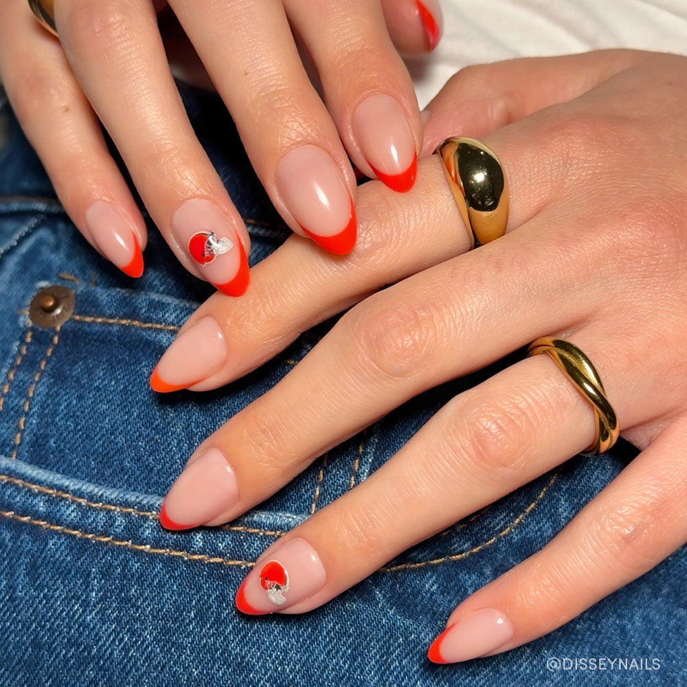 A close-up of hands wearing the glamnetic Cleveland Browns Press-On Nail Set, featuring almond-shaped nails with a nude base, bright red French tips, and a silver heart accent—perfect for game day. Gold rings and blue denim jeans complete the look.
