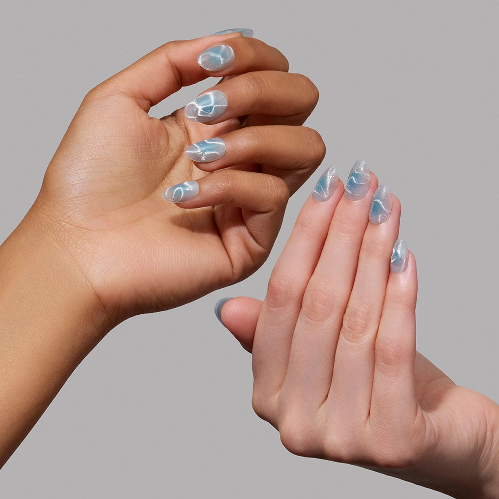 Two hands with different skin tones display manicured short oval nails painted with glamnetic’s Cloud Dance, a light blue polish featuring a white marbled, water-inspired pattern, set against a plain gray background. - Product image