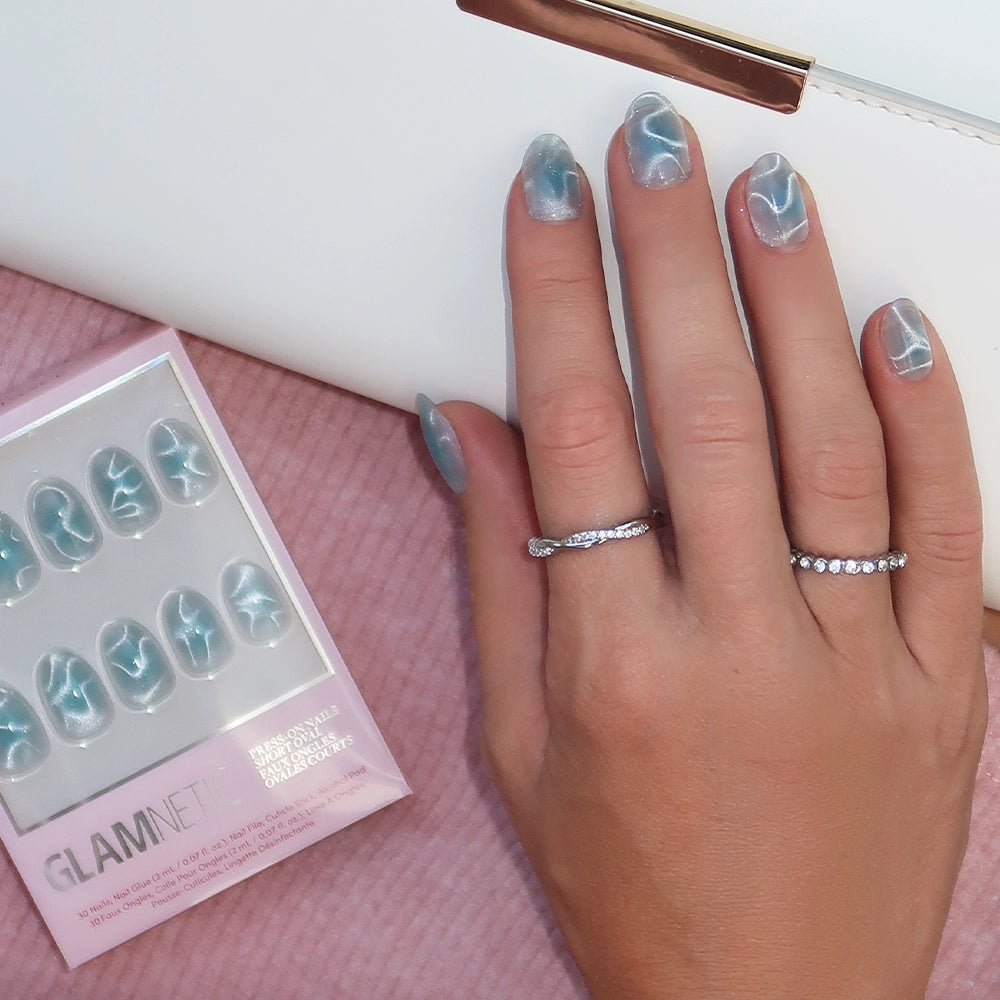 A hand with blue marble nail art and two rings rests on a white clutch, showcasing chic short oval nails, while a box of glamnetic Cloud Dance press-on nails lies nearby on a pink textured surface. - Product image