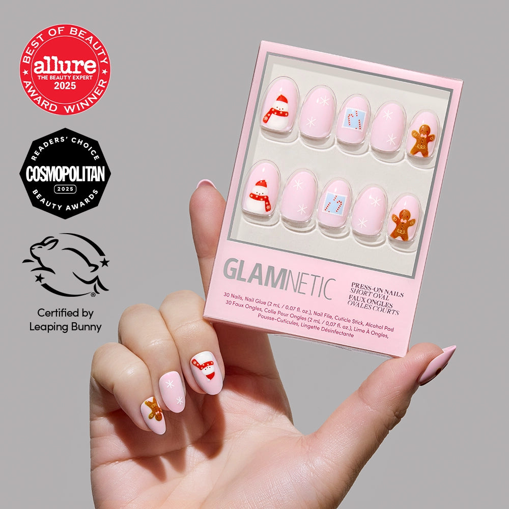 A hand holds a box of glamnetic Cocoa & Kisses holiday press-on nails, showcasing festive designs such as gingerbread men and candy canes. Beauty and cruelty-free awards are displayed to the left of the short oval pink nails against a neutral background. - Product image