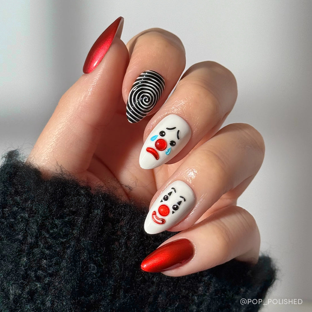 A hand with short almond nails wears glamnetics Crybaby Clown set: red chrome, black-and-white spiral, and white 3D clown nails with sad faces, all against a dark fuzzy sleeve.