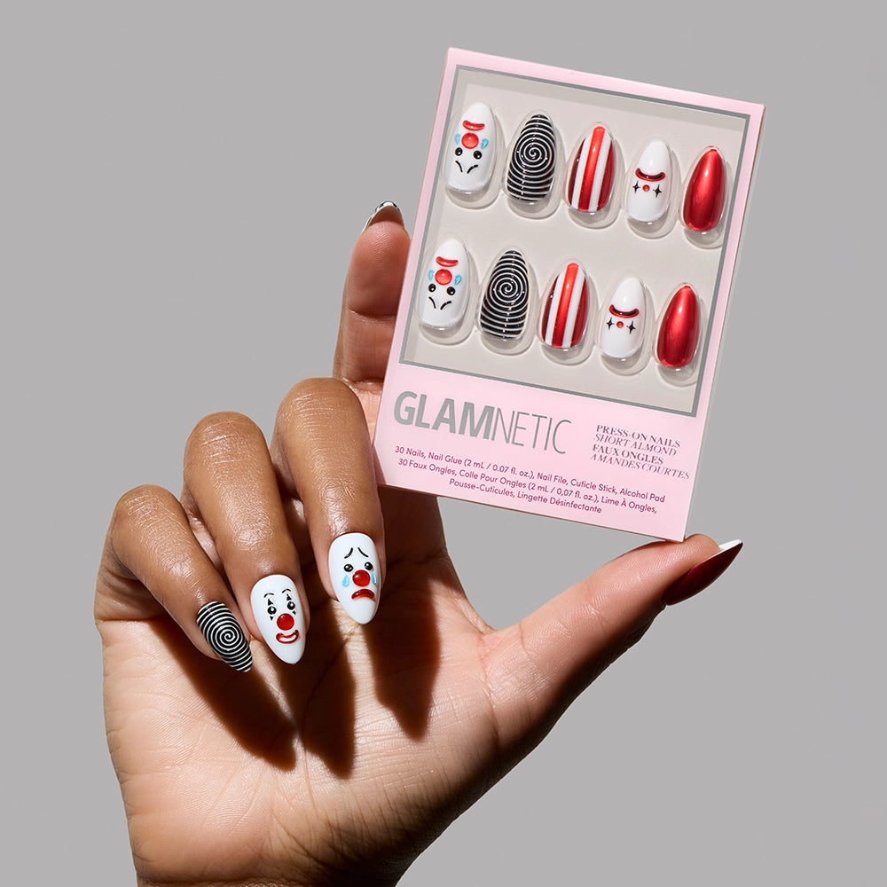 A hand with short almond nails featuring 3D clown art, red and white stripes, and spirals holds the glamnetic Crybaby Clown nail set with matching designs against a gray background.