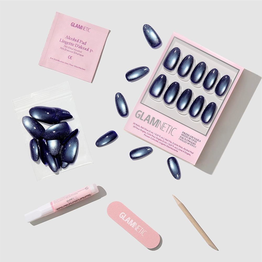 Glamnetic Crystal Cantation press-on nail kit featuring medium oval nails in a cat-eye style metallic blue with a luxurious velvet finish. The set includes a box of Crystal Cantation press-on nails, a plastic bag of spare nails, an alcohol pad, nail glue, a pink nail file, and a wooden cuticle stick on a light gray background. - Product image