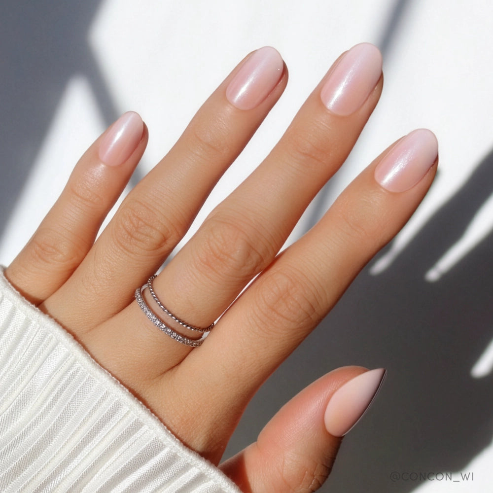 A hand with neatly manicured, short oval nails painted in glamnetic's Crystal Glaze, a soft pale pink. Two thin diamond-studded rings are stacked on the fingers, and the white ribbed sleeve of a sweater is visible. - Product image