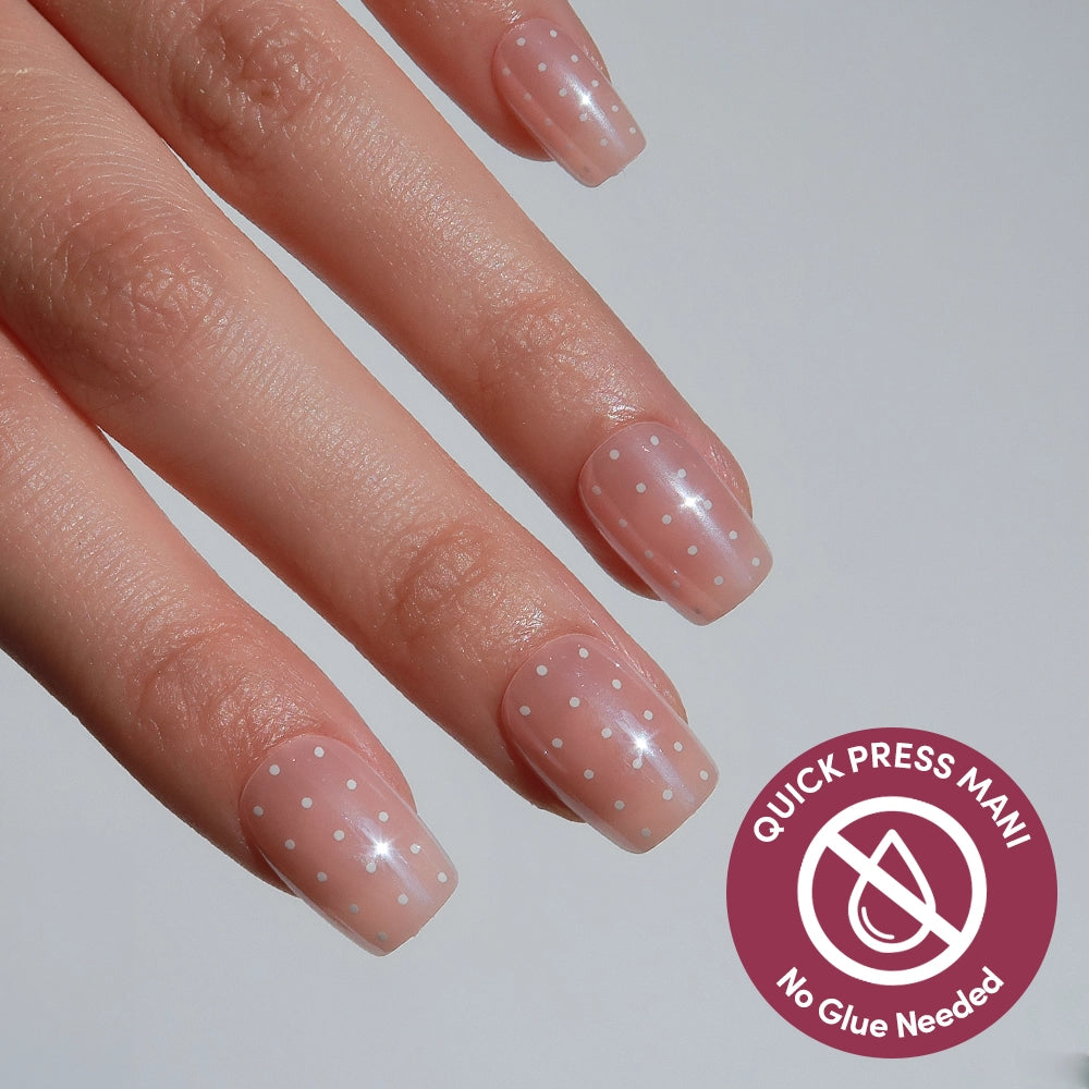 A hand with light pink, square-shaped Dainty Dots press-on nails by glamnetic, featuring small white polka dots. A circular badge reads Quick Press Mani, No Glue Needed.