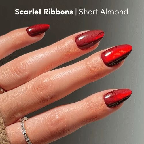A hand with short almond nails in glossy red and scarlet velvet, wearing a thin silver ring. Text: Scarlet Ribbons by glamnetic | Press-On Nail Set. - Product image