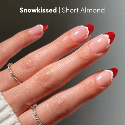 A hand with short almond-shaped nails showcases a red French tip and wavy white lines using the Snow Kissed set by glamnetic, paired with silver rings and a white sweater for a salon-quality look.
