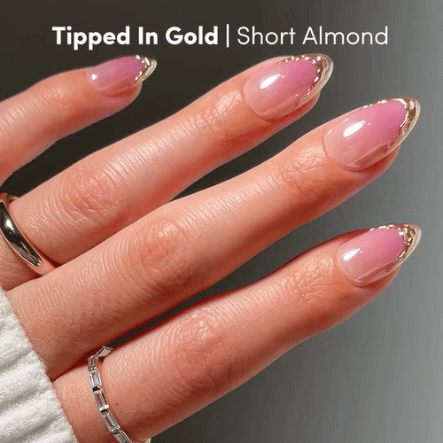 A hand with short almond nails in a pink base and gold French tips wears silver rings and a cream sweater. Text: Tipped in Gold by glamnetic | Short Almond.