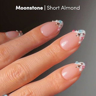 Close-up of a hand with short almond French tip nails featuring the "Moonstone" design by glamnetic, adorned with pastel beads, silver stars, and swirls on a pale nude base—ideal for achieving jewel-encrusted manicure elegance.