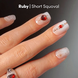 A hand with short, squoval nails showcases salon-quality details—white polish, ruby-red gem accents, gold studs, and small star designs—capturing the premium nail art of glamnetic Ruby Press-On Nails. A silver ring with a red gem sits on the index finger. Text reads: glamnetic | Ruby Press-On Nails | Short Squoval.
