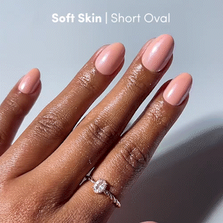 A close-up of a hand with a deep skin tone showcases short oval nails painted in glamnetic’s Soft Skin, a glossy soft pink polish that creates the perfect nude manicure. The fingers are moisturized, and one is adorned with a delicate twisted-band diamond ring. Text at the top reads: glamnetic | Soft Skin | Short Oval. - Product image