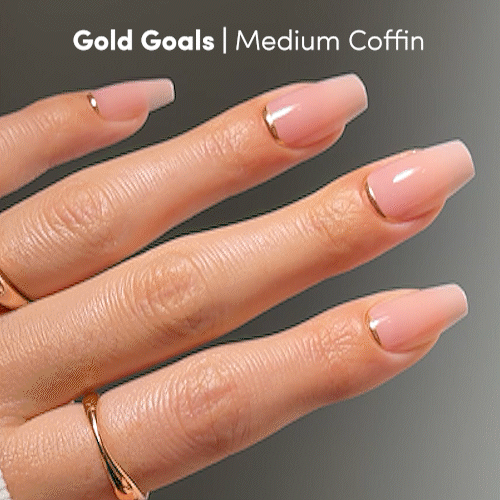 A hand with medium-length coffin-shaped nails, painted a soft pink shade with slim gold tips, showcases glamnetic’s Gold Goals Press-On Nails. The person accessorizes with gold rings for an extra touch of elegance. Text reads: Gold Goals | Medium Coffin.