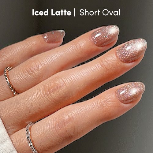 A hand displays glamnetic’s Iced Latte press-on nails in a short oval shape—a chic nude mani with a shimmery, iced latte hue. Two silver rings adorn the fingers, while “Iced Latte | Short Oval” appears in the top left corner. - Product image