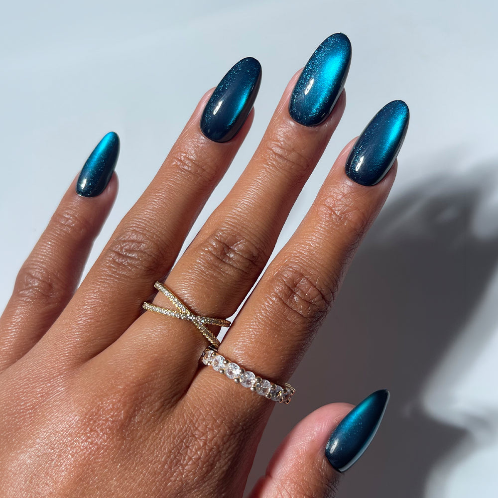 A hand displays almond-shaped nails adorned with glamnetic’s Dark Waters press-on set—a mesmerizing, metallic teal cat-eye finish that sparkles like gemstones. The look is accented by two gold rings: one with an x-shaped design, the other a slender band of clear stones. Ideal for those seeking long-lasting style without commitment, these quality press-ons offer effortless glamour. A soft, light gray background enhances the striking effect. - Product image