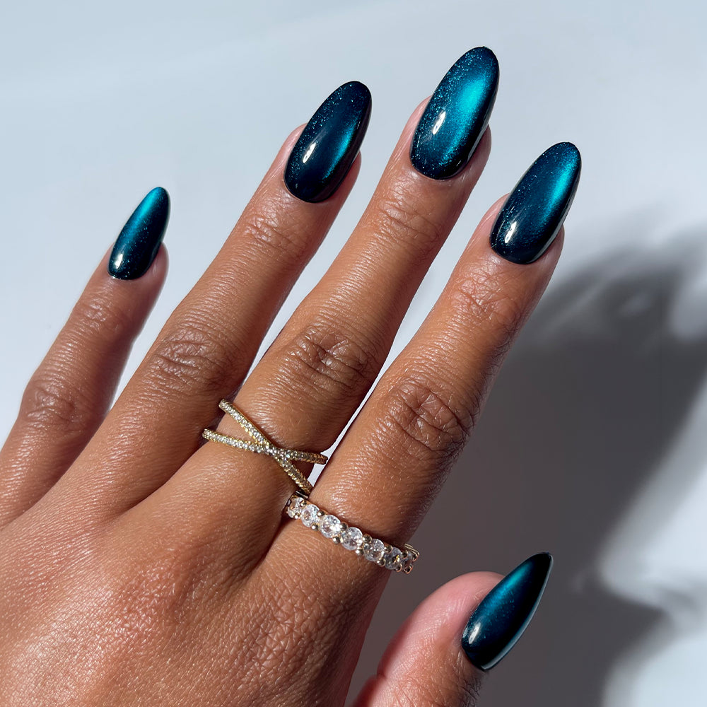 A hand displays almond-shaped nails adorned with glamnetic’s Dark Waters press-on set—a mesmerizing, metallic teal cat-eye finish that sparkles like gemstones. The look is accented by two gold rings: one with an x-shaped design, the other a slender band of clear stones. Ideal for those seeking long-lasting style without commitment, these quality press-ons offer effortless glamour. A soft, light gray background enhances the striking effect.