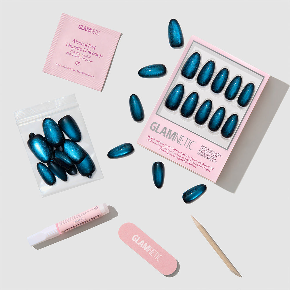 A set of glamnetic Dark Waters blue press-on nails, along with an alcohol pad, nail glue, nail file, wooden cuticle stick, and a zip bag, are arranged neatly on a white surface—an easy way to achieve long-lasting salon-quality nails at home. - Product image