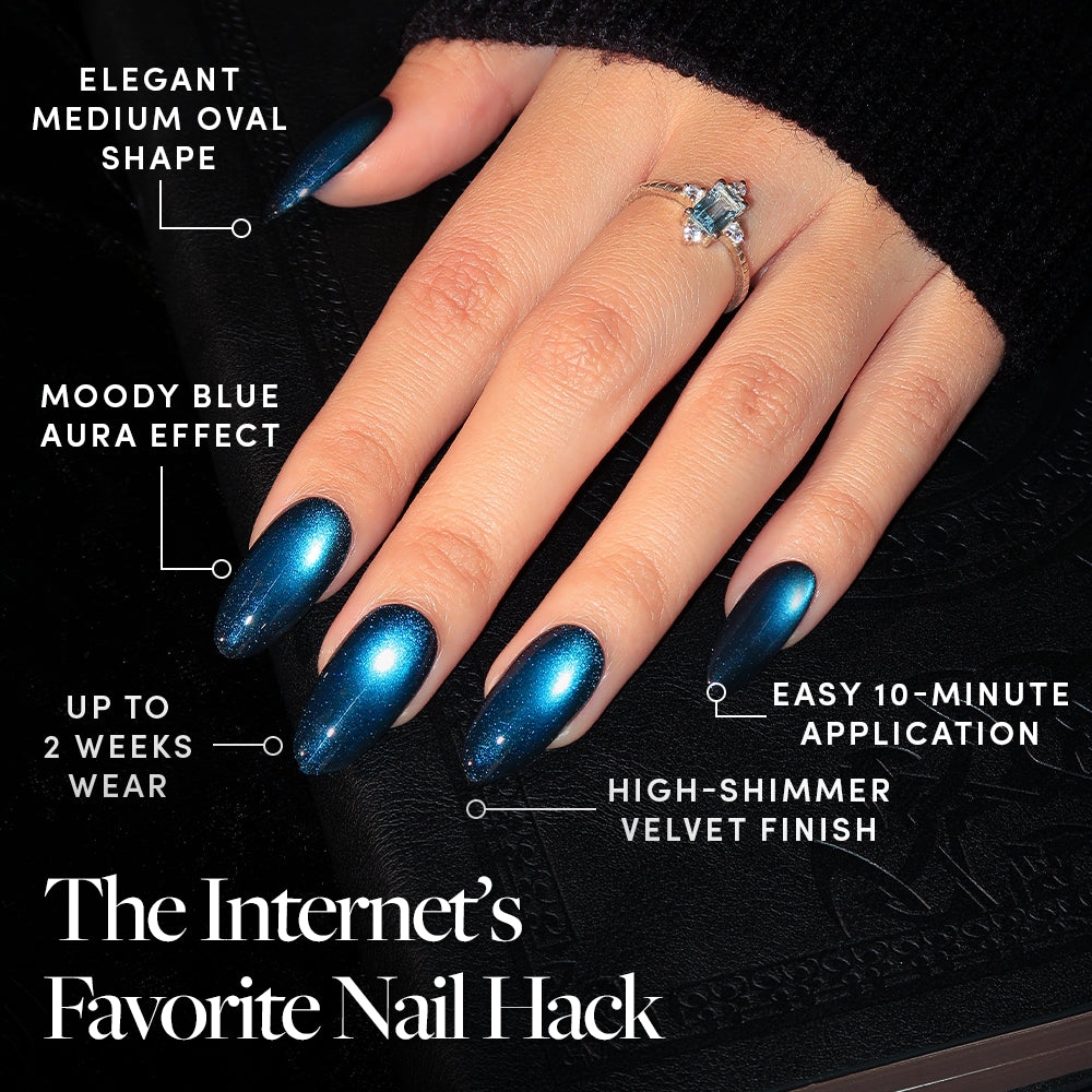 A hand adorned with glamnetic’s Dark Waters press-on nails—featuring elegant, medium oval-shaped tips in a moody blue shimmer and a luxurious velvet finish—wears a silver ring set with a blue gemstone. Text highlights include 10-minute application and up to 2 weeks of long-lasting wear. - Product image