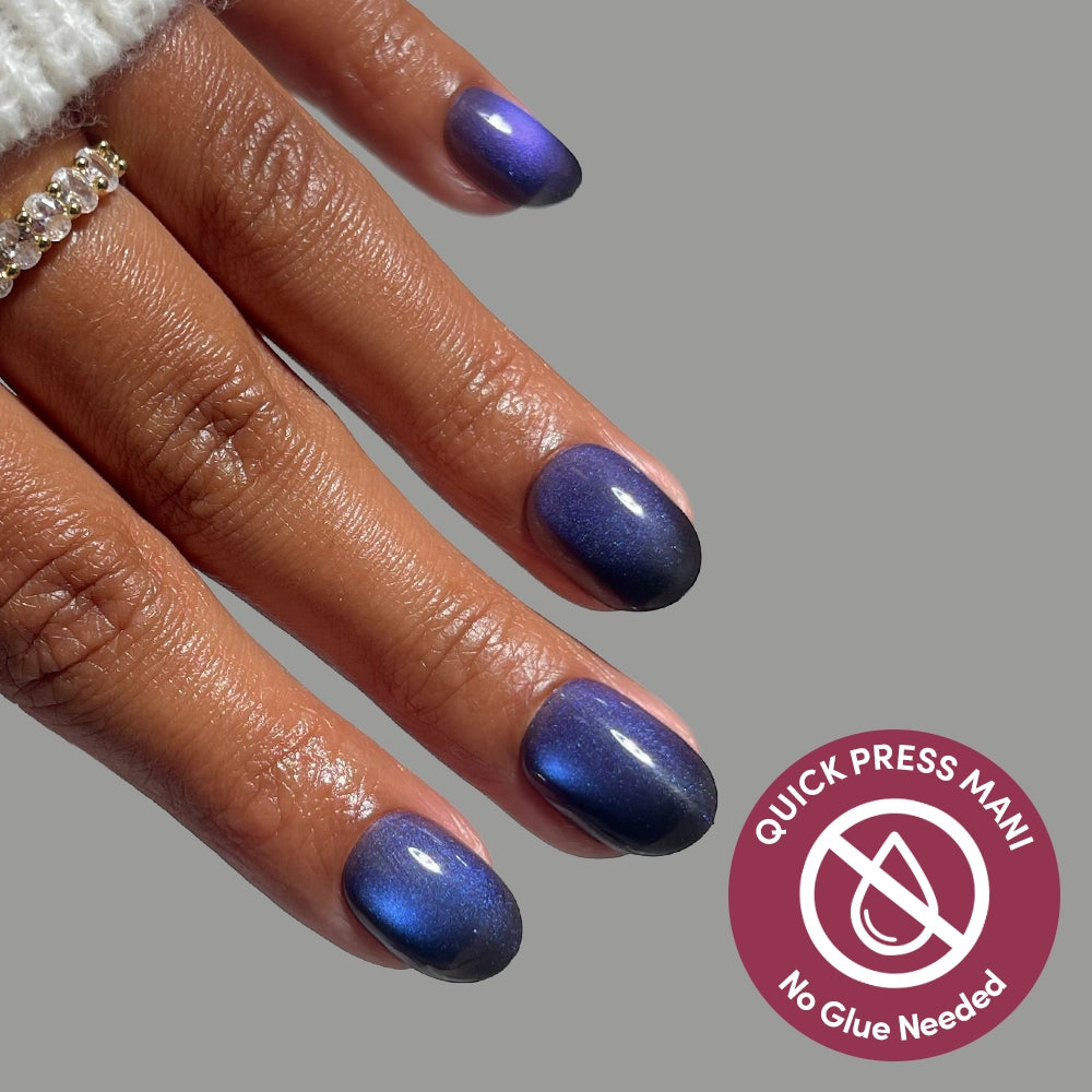 Hand with extra short round Deep Winter nails by glamnetic in a shimmering velvet purple polish, with a circular badge reading Quick Press Mani, No Glue Needed featuring a crossed-out water drop icon.