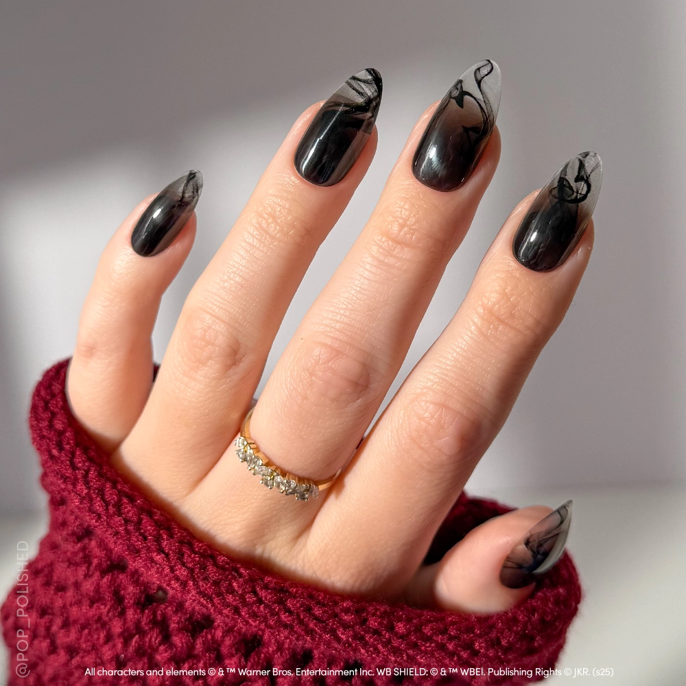 A hand with long, almond-shaped glamnetic Dementor™ press-on nails in black with smoky abstract tips, adorned with a gold ring and partially covered by a textured burgundy sleeve.