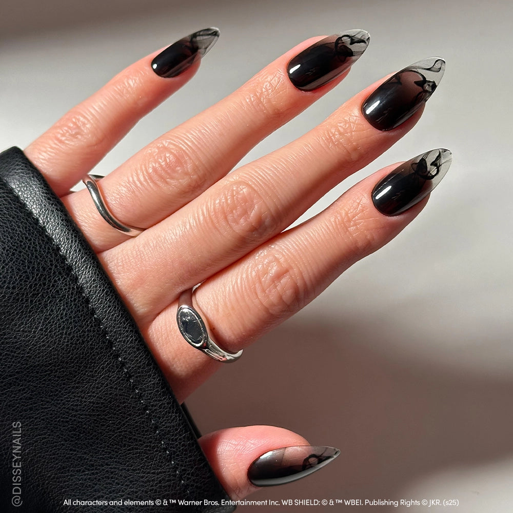 A hand with long almond-shaped nails from glamnetic's Dementor™ set features a black-to-clear gradient and glossy finish, adorned with silver rings and partially covered by black fabric in a well-lit image. - Product image