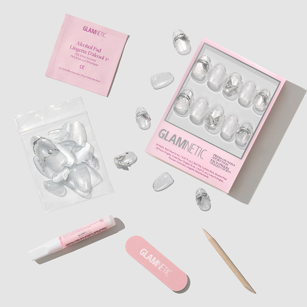 A flat lay of the glamnetic Diamond Press-On Nails kit showcases short oval silver glitter nails, a pink nail file, wooden cuticle stick, nail glue, alcohol pad, and extra nails on a light gray background. - Product image